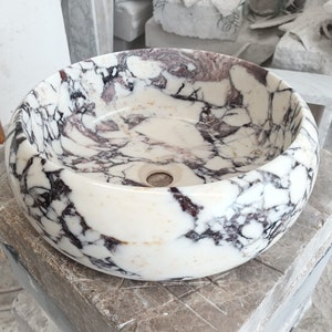 May include: A white marble sink with purple and black veining. The sink is round and has a smooth, polished finish.