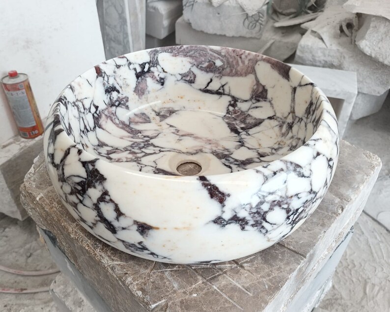May include: A white marble sink with purple and black veining. The sink is round and has a smooth, polished finish.