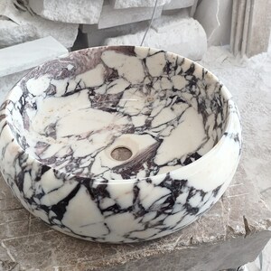 May include: A white marble sink with black and purple veining. The sink is round and has a smooth, polished finish.