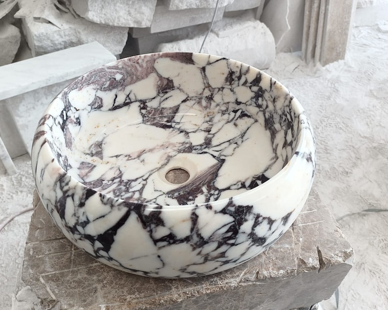May include: A white marble sink with black and purple veining. The sink is round and has a smooth, polished finish.
