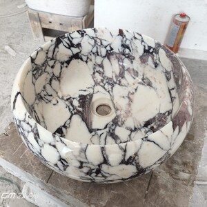 May include: A white marble sink with black and purple veining. The sink is round and has a smooth, polished finish.