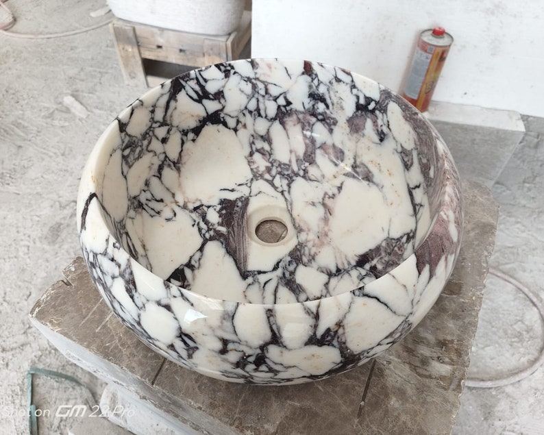 May include: A white marble sink with black and purple veining. The sink is round and has a smooth, polished finish.