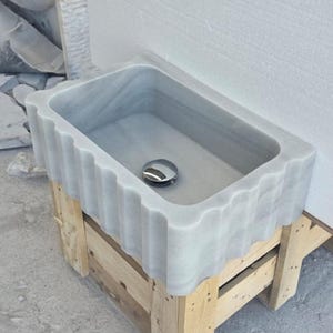 May include: A rectangular marble sink with a wavy, fluted edge. The sink is light gray with subtle veining and a polished chrome drain. It rests on a wooden pallet, showcasing a natural stone bathroom fixture.
