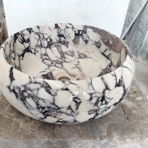 May include: A white marble sink with a round, bowl-shaped design. The sink has a swirling pattern of black and purple veins throughout the surface.