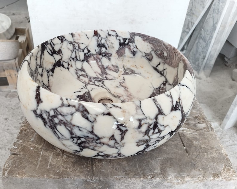 May include: A white marble sink with a round, bowl-shaped design. The sink has a swirling pattern of black and purple veins throughout the surface.