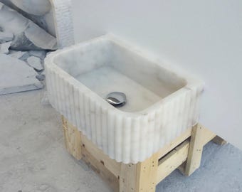 May include: A rectangular white marble sink with a fluted exterior design. The sink has a polished chrome drain and is set on a wooden support structure. The marble has subtle gray veining.