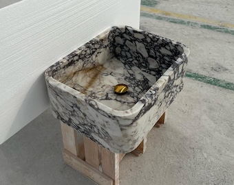 Puede incluir: Un lavabo de mármol blanco con vetas negras y marrones. El lavabo es rectangular y tiene un borde redondeado. Está sentado en un soporte de madera.