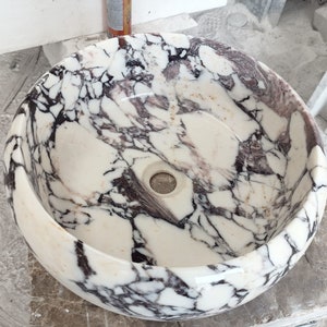 May include: A white marble sink with a swirling pattern of black and purple veins. The sink is round and has a smooth, polished finish.