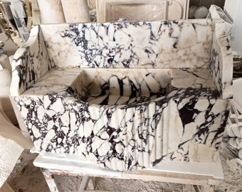 May include: A white marble sink with black veining, featuring a curved design and a raised backsplash. The sink is crafted from a single piece of marble and has a unique, organic look.