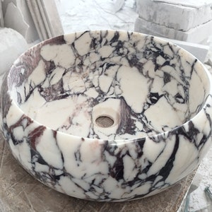 May include: A white marble sink with a round shape and a purple and black marbling pattern. The sink has a small drain hole in the center.