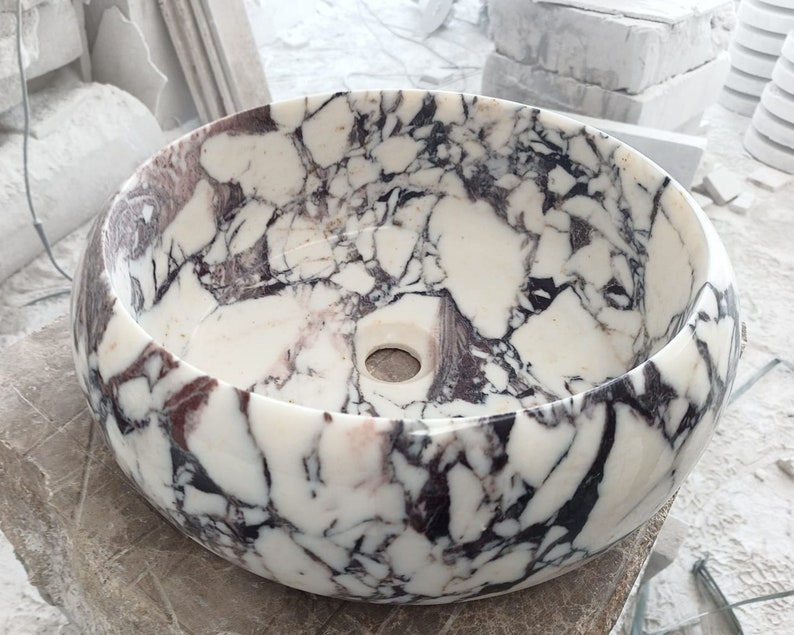 May include: A white marble sink with a round shape and a purple and black marbling pattern. The sink has a small drain hole in the center.