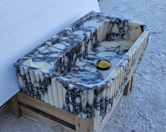 Puede incluir: Un lavabo rectangular de mármol con un patrón veteado blanco y gris oscuro. El lavabo tiene un exterior estriado y un desagüe dorado. Está colocado sobre un palet de madera.
