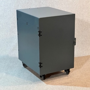 Op de afbeelding: Een grijze metalen kast met een deur en wielen. De kast heeft zwarte scharnieren en een slot.