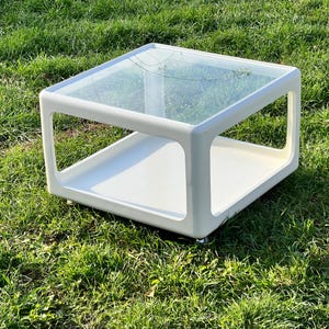 K&ouml;nnte beinhalten: Wei&szlig;er Couchtisch mit Glasplatte und einer unteren Ablage. Der Tisch hat ein modernes Design und steht auf einem gr&uuml;nen Rasen.