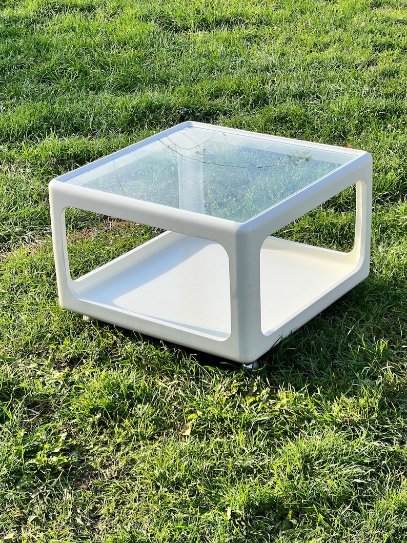 K&ouml;nnte beinhalten: Wei&szlig;er Couchtisch mit Glasplatte und einer unteren Ablage. Der Tisch hat ein modernes Design und steht auf einem gr&uuml;nen Rasen.