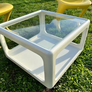 K&ouml;nnte beinhalten: Wei&szlig;er quadratischer Couchtisch mit Glasplatte und einer unteren Ablage. Der Tisch steht auf gr&uuml;nem Gras.
