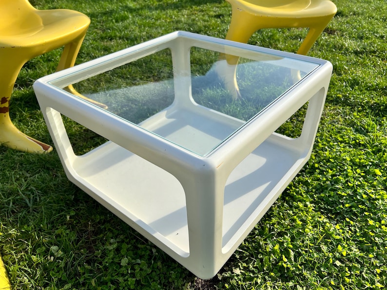 K&ouml;nnte beinhalten: Wei&szlig;er quadratischer Couchtisch mit Glasplatte und einer unteren Ablage. Der Tisch steht auf gr&uuml;nem Gras.