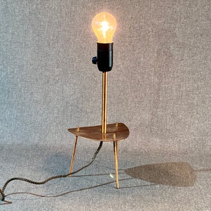 May include: A gold-toned table lamp with a black socket and a clear light bulb. The lamp has a triangular base with three legs.