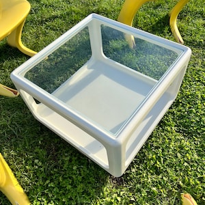 K&ouml;nnte beinhalten: Wei&szlig;er quadratischer Couchtisch mit Glasplatte. Der Tisch hat ein unteres Regal und steht auf gr&uuml;nem Gras.