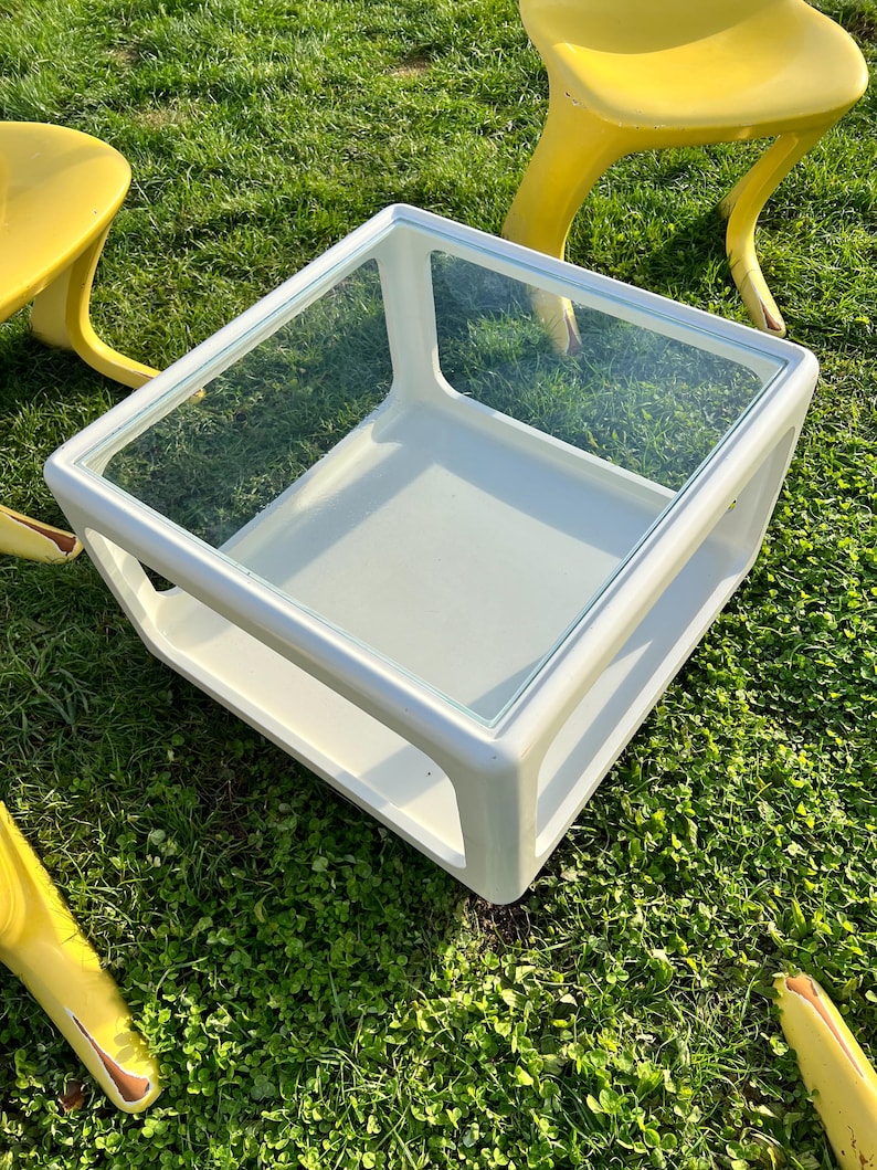 K&ouml;nnte beinhalten: Wei&szlig;er quadratischer Couchtisch mit Glasplatte. Der Tisch hat ein unteres Regal und steht auf gr&uuml;nem Gras.