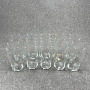 Puede incluir: Un conjunto de vasos transparentes sin pie. Los vasos tienen una base redondeada y una parte superior ligeramente ensanchada. Están dispuestos en filas sobre una superficie gris. Los vasos están vacíos y parecen ser de vidrio.