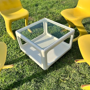 K&ouml;nnte beinhalten: Ein wei&szlig;er quadratischer Couchtisch mit Glasplatte steht auf einem gr&uuml;nen Rasen, umgeben von vier gelben Plastikst&uuml;hlen.