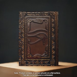 May include: A brown leather-bound journal with a gold embossed design featuring the Eye of Horus. The journal has a decorative border with intricate patterns.