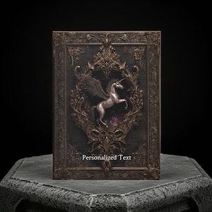 May include: A dark, ornate book with a silver winged unicorn design. The cover features intricate gold filigree and the text "Personalized Text". The book is displayed on a textured stone surface against a black background.