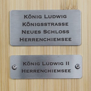 May include: Two silver metal plaques with black text. The top plaque reads "König Ludwig, Königstrasse, Neues Schloss, Herrenchiemsee". The bottom plaque reads "König Ludwig II, Herrenchiemsee".