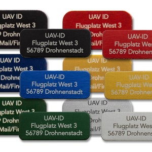 Puede incluir: Nueve etiquetas rectangulares de metal con esquinas redondeadas en varios colores, incluyendo negro, azul, rojo, dorado, verde y plateado. Cada etiqueta tiene texto blanco que dice "UAV-ID Flugplatz West 3 56789 Drohnenstadt" o una variación similar.