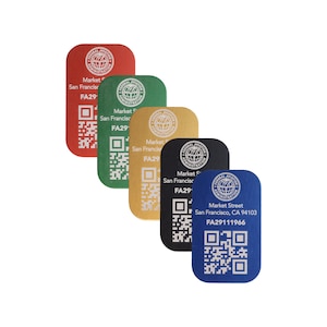 Op de afbeelding: Vijf metalen tags in blauw, zwart, goud, groen en rood. Elke tag heeft een cirkelvormig logo met de tekst "Federal Aviation Administration" en een wereldbeeld. De tags bevatten ook de tekst "Market Street", "San Francisco" en een uniek identificatienummer. Elke tag heeft een QR-code.