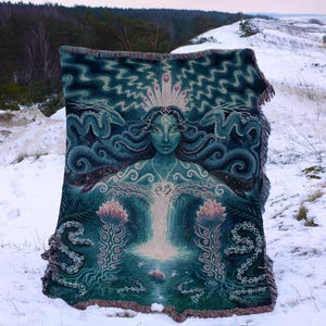 May include: A teal and green tapestry featuring a central female figure with flowing hair and a crown. The design includes floral and serpent motifs. The tapestry is displayed outdoors in a snowy landscape.