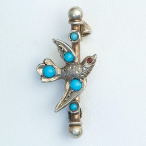 May include: A silver-toned brooch featuring a bird design with turquoise and red accents. The bird is perched on a vertical bar with turquoise beads. The brooch has a detailed, vintage aesthetic.