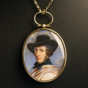 May include: An oval gold pendant necklace featuring a miniature portrait painting of a person wearing a dark hat, a black necktie, and a striped shawl against a blue sky background. The pendant is suspended from a delicate gold chain link.