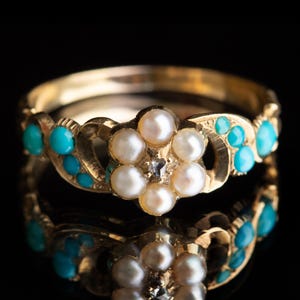 Puede incluir: Un anillo de oro con un diseño floral, perlas blancas centrales y un pequeño diamante. La banda está adornada con piedras turquesas. El anillo está sobre un fondo negro, con su reflejo visible.