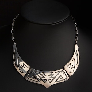 May include: A silver-toned statement necklace with a geometric design. The necklace features a curved bib-style pendant with cut-out patterns and is connected to a chain. The design includes angular shapes and lines, creating a bold and artistic look.