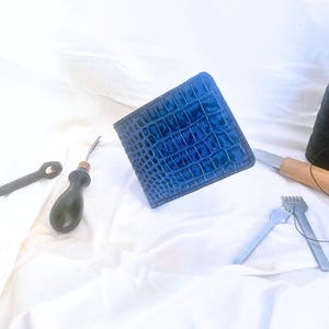 Może przedstawiać: Niebieski skórzany portfel z wzorem krokodyla i narzędziami do obróbki skóry. Narzędzia obejmują nóż, szpulę czarnej nici i różne metalowe narzędzia. Portfel ma około 11,4 cm szerokości i 8,9 cm wysokości.