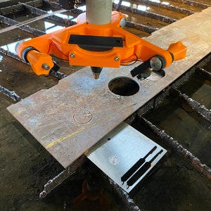 Op de afbeelding: Een close-up van een oranje metalen snijgereedschap met een zwarte handgreep, gemonteerd op een metalen arm. Het gereedschap is gepositioneerd boven een metalen plaat met verschillende uitgesneden vormen, waaronder een cirkel en twee lange, dunne rechthoeken. De metalen plaat ligt op een metalen rooster.