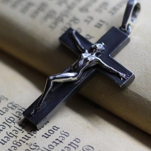 May include: A dark metallic cross pendant featuring a polished silver-toned figure attached to the front. The cross has a smooth, matte finish and includes a small metal loop at the top for attachment to a chain or cord, presented as a jewelry item.