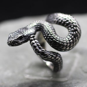 May include: A silver snake ring with a detailed, textured design. The snake is coiled and its head is raised, giving the ring a dramatic look.