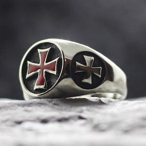Könnte beinhalten: Ein silberfarbener Metallring mit einer kreisförmigen Vorderseite, die ein rotes Emaille-Kreuz auf dunklem Hintergrund zeigt. Ein kleineres, schlichtes silbernes Kreuz ist an der Seite des Rings eingeprägt. Der Ring hat eine polierte Oberfläche.
