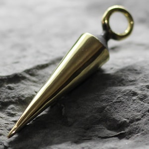 May include: A gold-colored metal pendant shaped like a cone with a small ring at the top. The pendant is sitting on a dark gray textured surface.