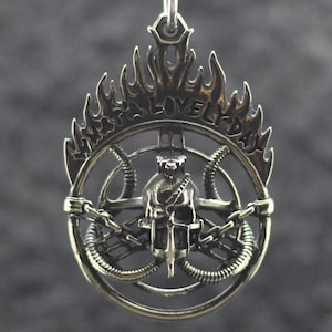 May include: A silver pendant on a chain featuring a central skull design surrounded by a circular frame with mechanical tubes and flames. The text WHAT A LOVELY DAY is engraved along the top edge of the flames. The metal has a polished finish.