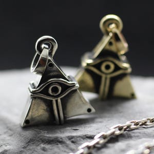 May include: Two pyramid-shaped pendants featuring an engraved eye symbol on the front face. One pendant is finished in a polished silver tone, while the other has a gold tone. Each piece includes a circular bail at the top for a chain attachment.