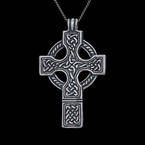 May include: A silver-toned metal Celtic cross pendant suspended from a matching chain. The cross features intricate, raised knotwork patterns throughout its surface, centered within a circular ring that also displays a twisted rope-like texture.