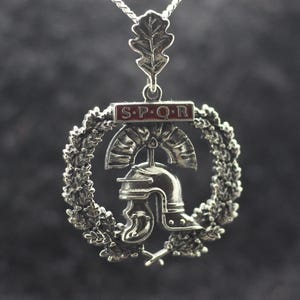 May include: A silver-toned pendant featuring a Roman galea helmet centered within a circular wreath of oak leaves. Above the helmet is a rectangular bar with the red-lettered text S.P.Q.R. The pendant hangs from a silver chain by an oak leaf bail.