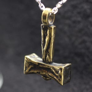 May include: A metallic pendant shaped like a hammer with a textured, weathered gold-tone finish. The pendant features a vertical handle and a wide, horizontal head, suspended from a silver-colored metal chain link necklace.