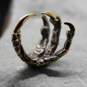 Op de afbeelding: Een metalen ring in de vorm van een vogelklauw met een donkere, geoxideerde afwerking en gepolijste, goudkleurige punten. De band heeft een gestructureerd, schubachtig ontwerp dat het uiterlijk van een roofvogelpoot nabootst in een open cirkelvorm.