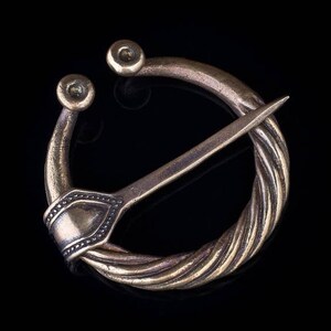May include: A bronze-toned annular brooch featuring a twisted metal ring design with two rounded terminals at the top opening. A long, pointed pin extends across the center, secured by a smooth, flat metal plate with small decorative dot detailing.