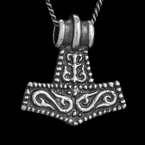 May include: A silver-toned metal pendant shaped like a hammer, suspended from a matching silver-toned chain. The pendant features intricate, embossed scrollwork and geometric patterns across its surface, with a prominent bail at the top.